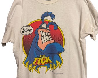 May include: White t-shirt featuring a cartoon character, The Tick, with a blue costume and a large grin. The character is set against a red and yellow background, with the words "EVIL BEWARE!!" and "THE TICK" printed on the shirt.