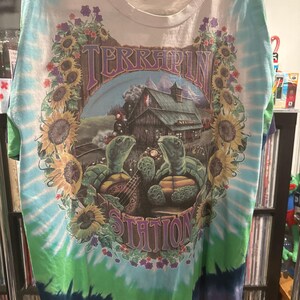 May include: A tie-dye t-shirt with a graphic featuring two turtles playing instruments in front of a cabin. The shirt has the words "TERRAPIN STATION" and is surrounded by sunflowers and purple flowers.