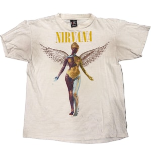 Puede incluir: Una camiseta de manga corta de color crema con la palabra NIRVANA en texto amarillo sobre un gráfico de una figura alada. La figura se muestra con un lado que revela la estructura anatómica muscular y ósea en tonos dorados y azules.