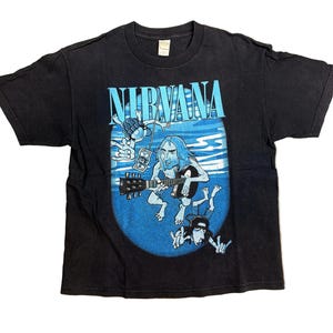 以下が含まれることがあります： 黒いTシャツ。青色のブロック文字で「NIRVANA」のバンド名が描かれ、その下に漫画のグラフィックがあります。グラフィックは、ギターを演奏するミュージシャンと、青い楕円の中にいる他の漫画のキャラクターを描いています。