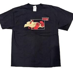 May include: Black t-shirt featuring a graphic of the Hellboy character in red, with a white cape and a large red fist. The word "Hellboy" is printed in red above the graphic. The shirt is made of a soft material.