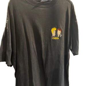 Op de afbeelding: Donkergrijs T-shirt met een geborduurde afbeelding van twee stripfiguren met geel en bruin haar. De afbeelding bevat de tekst "Beavis and Butt-Head". Het T-shirt hangt aan een grijze hanger.