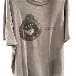 May include: A grey t-shirt with a circular graphic of a monkey skull wearing a halo. The band name "Pixies" is printed in a rectangular box. The t-shirt is hanging on a hanger. The fabric appears to be cotton.