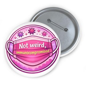 May include: A white button badge with a pink and purple design. The design features a pink face mask with the words "Not weird, immunocompromised" in white text. The badge also includes illustrations of viruses and stars.