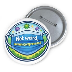 May include: A white button badge featuring a blue surgical mask design with the text "Not weird, immunocompromised." The design includes green and purple virus illustrations. A second, silver-coloured badge is partially visible.