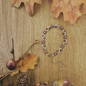 May include: A beaded bracelet featuring a repeating floral pattern made of small, dark brown and translucent white beads. The bracelet includes a metallic lobster claw clasp and an adjustable chain extension with a small, round metal charm at the end.