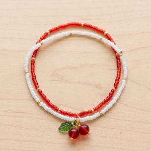 May include: A double-strand beaded bracelet featuring one strand of small red beads and one strand of small white beads, accented with gold-toned spacer beads. A dangling charm of two red spherical beads and a green leaf is attached to the center.