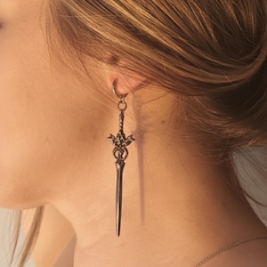 May include: A silver-colored sword-shaped earring with intricate detailing hangs from a hoop earring. The earring is suspended near the ear and neck of a person. A silver necklace is also visible.