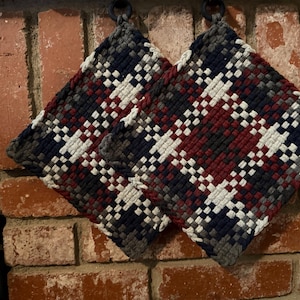 May include: Two square potholders featuring a woven plaid pattern in navy blue, deep red, white, and grey. Each potholder is constructed with a thick, textured weave and includes a small black circular loop at the top corner for convenient hanging.
