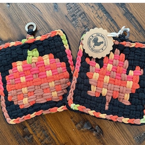 May include: Two square woven potholders featuring a black background with a pumpkin design on the left and a maple leaf design on the right. Both are crafted with red, orange, and yellow yarn, finished with a braided border and a metal ring.