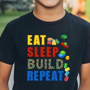 May include: Black t-shirt featuring the phrase "EAT SLEEP BUILD REPEAT" in bright block letters. The word "BUILD" is made of a mosaic of small blocks. Scattered around the text are various colourful toy blocks.