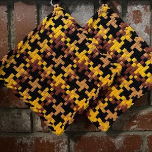 May include: Two square potholders featuring a woven, checkered pattern in vibrant yellow, deep black, and warm brown yarn. The potholders are constructed with a thick, textured loop design and are displayed in a diamond orientation.