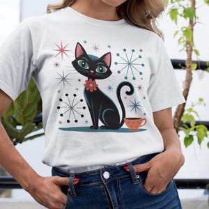 Könnte beinhalten: Weißes T-Shirt mit einer Cartoon-Katze mit rotem Blumenkragen, neben einer orangefarbenen Tasse. Das Design umfasst sternförmige und schneeflockenartige Formen in verschiedenen Farben. Die Katze hat grüne Augen und einen eingerollten Schwanz.