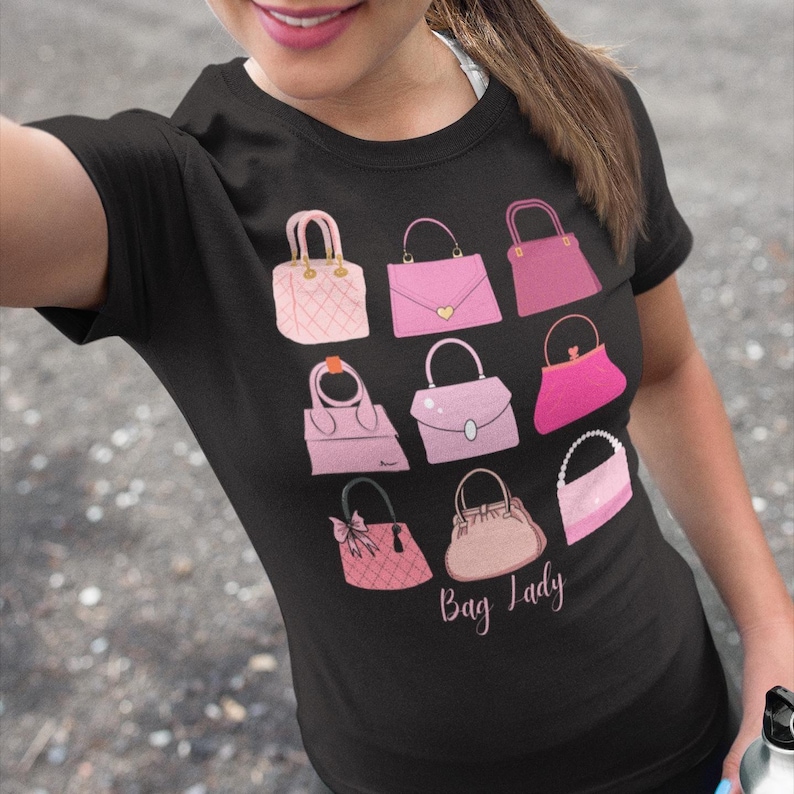 May include: Black t-shirt featuring a collection of pink handbags in various styles. The design includes a quilted bag, a heart-shaped clasp bag, and a bag with a bow. The words "Bag Lady" are written in cursive.