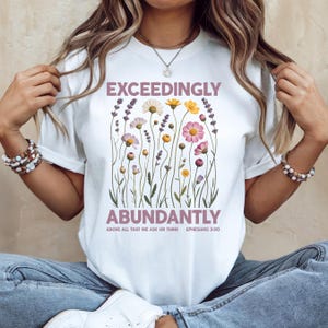 May include: White t-shirt with the words "EXCEEDINGLY ABUNDANTLY" in a mauve color, with a floral design of various flowers in shades of pink, yellow, and white. The text "ABOVE ALL THAT WE ASK OR THINK EPHESIANS 3:20" is below the main text.