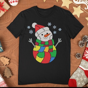 Könnte beinhalten: Schwarzes T-Shirt mit einem bunten Schneemann-Design. Der Schneemann hat einen roten Hut, eine orangefarbene Nase und einen Patchwork-Schal und -Körper. Das Design beinhaltet Schneeflocken.