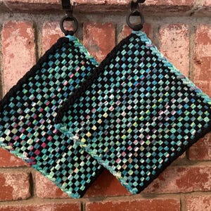 May include: Two square potholders featuring a woven checkerboard pattern in black, teal, light blue, and white. Each potholder has a solid black border and a black circular loop at the top corner for hanging, showcasing a dense, textured fabric construction.