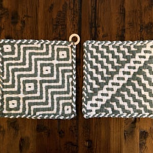 May include: Two square potholders made of woven cord in sage green and white. The left potholder features a geometric maze pattern, while the right one displays a diagonal chevron weave. Each piece includes a small wooden ring for hanging.