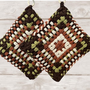 May include: Two square potholders featuring a woven geometric pattern in dark brown, light green, cream, and rust orange. Each potholder is constructed with a thick, textured fabric weave and includes a small metal ring at the top corner for hanging.