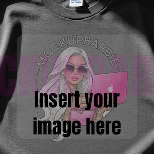 May include: Dark gray t-shirt with a graphic of a woman with sunglasses holding a pink laptop. The text "MOCKUPBARBIE" is in a circle above the woman, and "Insert your image here" is below. The shirt is on a wooden surface.