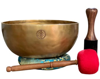 Puede incluir: Un cuenco tibetano dorado con un emblema decorativo se muestra sobre un cojín estampado. Se incluye una maza de madera con cabeza de fieltro rojo y un percutor de madera. El conjunto es una herramienta de meditación.