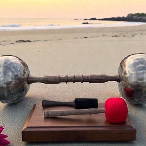May include: A large, silver singing bowl with a wooden handle and two mallets, one with a red head, on a wooden base. The bowl is on a sandy beach with the sea in the background.