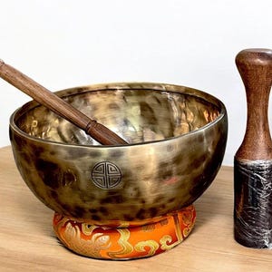 May include: A bronze singing bowl with a wooden mallet and a wooden striker. The bowl sits on an orange and gold patterned cushion. The mallet and striker are made of dark wood.