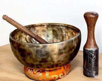 May include: A bronze singing bowl with a wooden mallet and a wooden striker. The bowl rests on an orange and gold patterned cushion. The mallet and striker are made of dark wood.