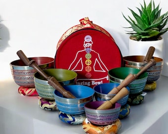May include: A collection of singing bowls in various colors, including blue, green, and red, with wooden strikers. The bowls are displayed on decorative cushions. A red carrying case with a chakra diagram is in the background.