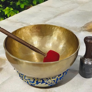 Puede incluir: Un cuenco tibetano dorado con una maza de madera y un percutor de fieltro rojo. El cuenco descansa sobre un cojín azul y dorado estampado. También se incluye un percutor de madera oscura.