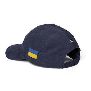 Puede incluir: Una gorra de béisbol de pana azul marino con un pequeño parche rectangular de la bandera de Ucrania en el lateral y las letras JB bordadas en blanco en el panel trasero. La gorra tiene una visera curva y un cierre de correa de tela.
