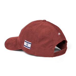 Peut inclure: Une casquette de baseball en velours côtelé marron avec un petit écusson rectangulaire du drapeau israélien sur le côté et les lettres JB brodées en blanc sur le panneau arrière. La casquette possède une visière courbée et une attache.