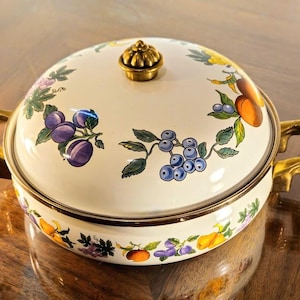 Puede incluir: Cazuela redonda de color crema con tapa, adornada con un diseño de frutas y flores. Cuenta con asas doradas y un pomo dorado en la tapa. El diseño incluye ciruelas, arándanos y albaricoques.