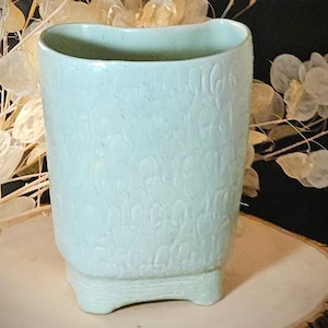 Peut inclure: Un vase en céramique vert clair avec un motif texturé. Le vase a une base rectangulaire avec de petits pieds et un dessus incurvé. Le fond comprend des plantes séchées et une tranche de bois.