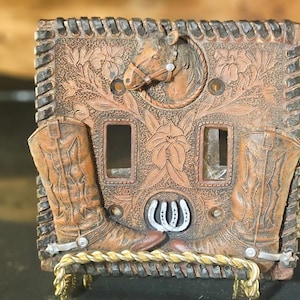 Könnte beinhalten: Eine dekorative Lichtschalterabdeckung mit Western-Thema. Die braune Abdeckung zeigt einen Pferdekopf, Cowboy-Stiefel und ein Hufeisen. Die Kanten sind mit einem geflochtenen Design strukturiert. Die Abdeckung wird von einem goldfarbenen Ständer getragen.