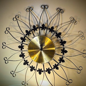 Peut inclure: Une horloge murale décorative avec un cadran doré et un cadre en métal noir. Le cadre présente un motif de soleil avec des accents de feuilles. Des chiffres romains marquent les heures sur le cadran.