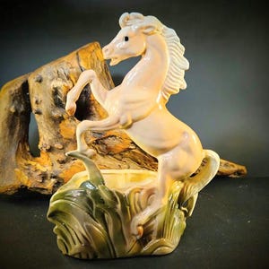 Op de afbeelding: Een crème-kleurige keramische paardenfiguur in een steigerende pose, met een witte manen en staart. Het paard staat op een voetstuk met groen en bruin gebladerte. De achtergrond bevat een stuk drijfhout.