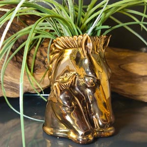 May include: A gold-coloured ceramic vase shaped like two horse heads. The vase is filled with green, grass-like plants. The vase has a glossy finish and is set against a blurred background of wood.