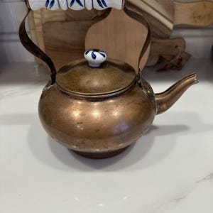 May include: A copper tea kettle with a rounded body and a curved spout. The handle features a white ceramic grip with blue floral patterns, and the lid is topped with a matching white and blue ceramic knob. The metal surface shows a patina.
