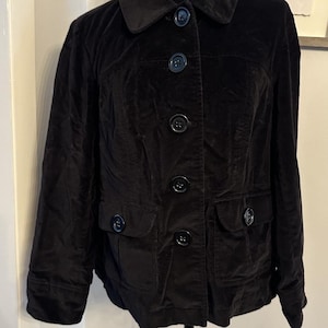 May include: A black velvet jacket featuring a classic collar, long sleeves, and a front button fastening. The garment includes two front pockets with buttoned flaps, and the overall design is finished with matching black buttons down the centre.