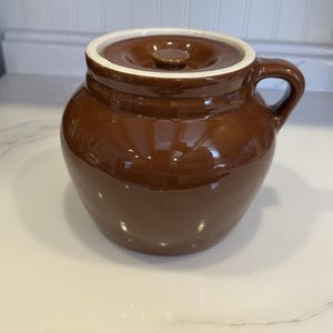Może przedstawiać: Błyszczący brązowy ceramiczny garnek z pasującą pokrywką i pojedynczym uchwytem z boku. Pojemnik ma zaokrąglony korpus i białą wewnętrzną krawędź, prezentując gładkie, odblaskowe wykończenie na zewnętrznej powierzchni naczynia z kamionki.