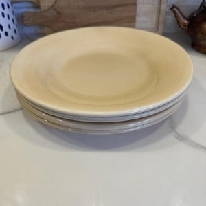 May include: Four beige ceramic plates stacked on a white surface. The plates have a shallow, rounded design. The plates are a solid colour with a glossy finish. Other kitchen items are visible in the background.