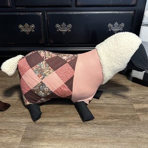 May include: A sheep-shaped decorative pillow with a patchwork quilt design on its body. The quilt features a mix of floral and geometric patterns in shades of pink and brown. The head and tail are white, and the legs are black.