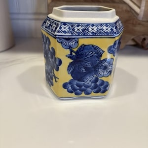 Pode incluir: Um vaso de cerâmica amarelo e azul com formato hexagonal. O vaso apresenta desenhos florais e de frutas azuis sobre um fundo amarelo. A borda superior é branca com um padrão geométrico azul.