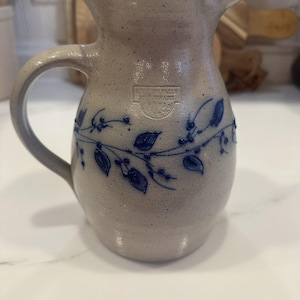 Puede incluir: Una jarra de gres beige con acabado moteado y un patrón de enredadera pintado en azul con hojas y bayas. La jarra tiene un asa curva y una marca estampada que dice Salmon Falls Stoneware 1993, Maine.