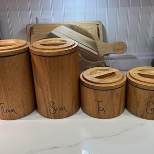 Puede incluir: Un juego de cuatro recipientes cilíndricos de madera con tapas a juego, que presentan las palabras grabadas Flour, Sugar, Tea y Coffee en la parte frontal. Los dos recipientes más altos están a la izquierda y los dos más bajos a la derecha.