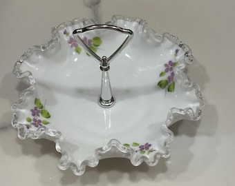 Puede incluir: Plato para servir blanco con borde con volantes y asa plateada. El plato presenta flores moradas pintadas a mano con hojas verdes. El asa es plateada y el plato está hecho de un material translúcido.