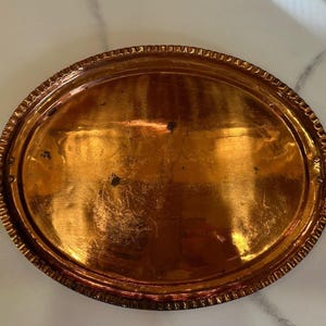 May include: Oval copper serving tray with two handles. The tray has a decorative, textured rim and a polished, reflective surface. The handles are a darker, contrasting metal. A vintage piece for serving or display.