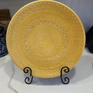 May include: A mustard-yellow ceramic plate with an embossed geometric pattern. The plate features concentric circles of diamond and dot designs. It is displayed on a black metal plate stand, showcasing its decorative details and craftsmanship.