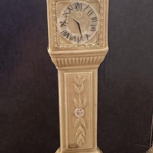 May include: A beige ceramic grandfather clock figurine with a clock face and Roman numerals. Two small animal figures sit atop the clock. The clock has decorative floral and leaf patterns.